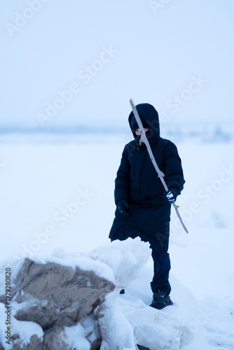 A boy digs through snow or ice with a stick. A teenager, holding a stick, walks along the frozen seashore on a frosty, cloudy winter day, checking the density of the snow along the way. 