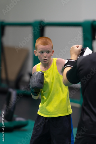A red-haired teenager practices punches on boxing mitts. The boy trains with a trainer. The young man practices MMA or boxing punches. High quality photo