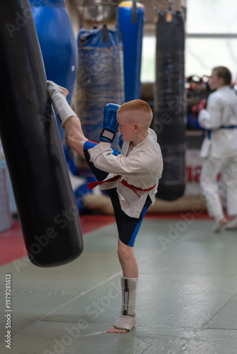 A teenager in a kimono jacket and shorts trains with a punching bag in a gym. Kickboxers train and exercise for competitions, tournaments, or events at a sports club. High quality photo
