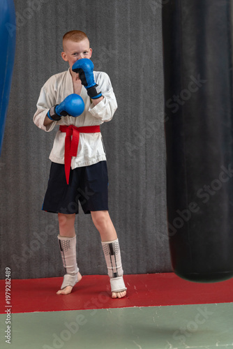Portrait of a red-haired smiling teenager wearing a kimono jacket, shorts, gloves and shin guards standing on a tatami mat in a gym. High quality photo