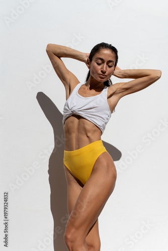 Obraz na płótnie Athletic adult woman in yellow bikini bottom and white sports top posing with ra