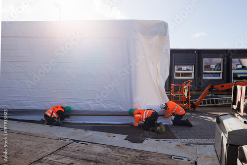 Three construction workers in full safety gear collaborating to wrap a prefabricated office unit, captured with sunlight flaring into the lens.