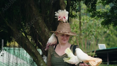 Young woman with hat holding colorful pigeons on hands and shoulders in tropical park on Koh Samui Thailand