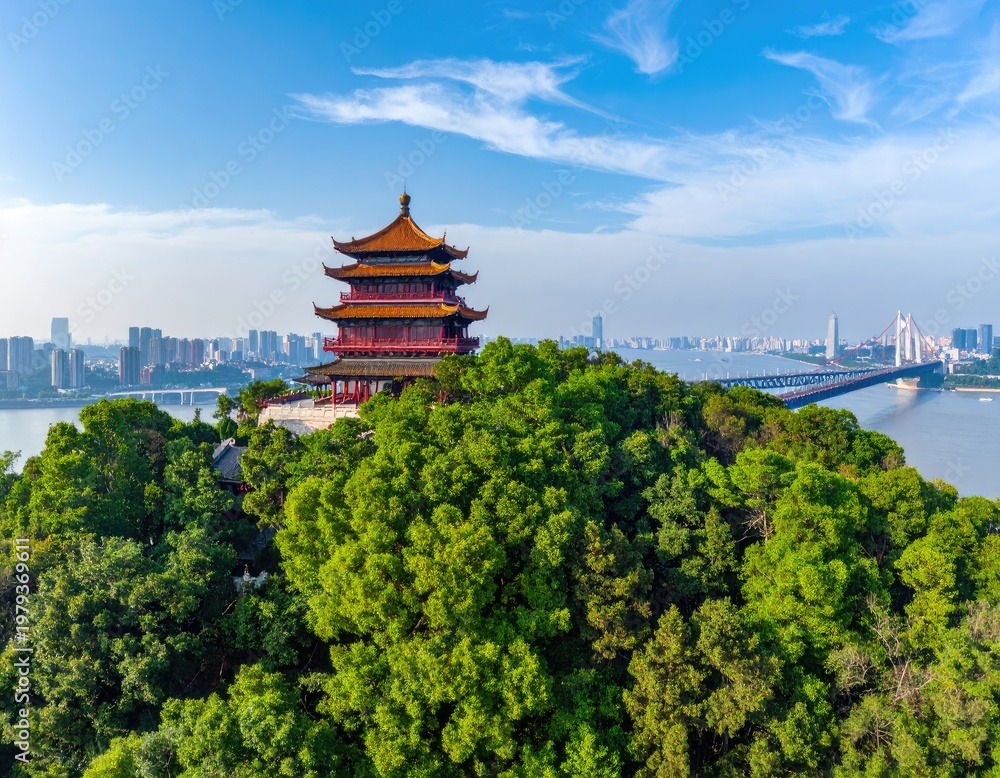 Obraz premium An aerial view showcases a traditional Chinese pagoda perched atop a lush green hill, overlooking a sprawling cityscape and a modern suspension bridge against a vibrant blue sky.