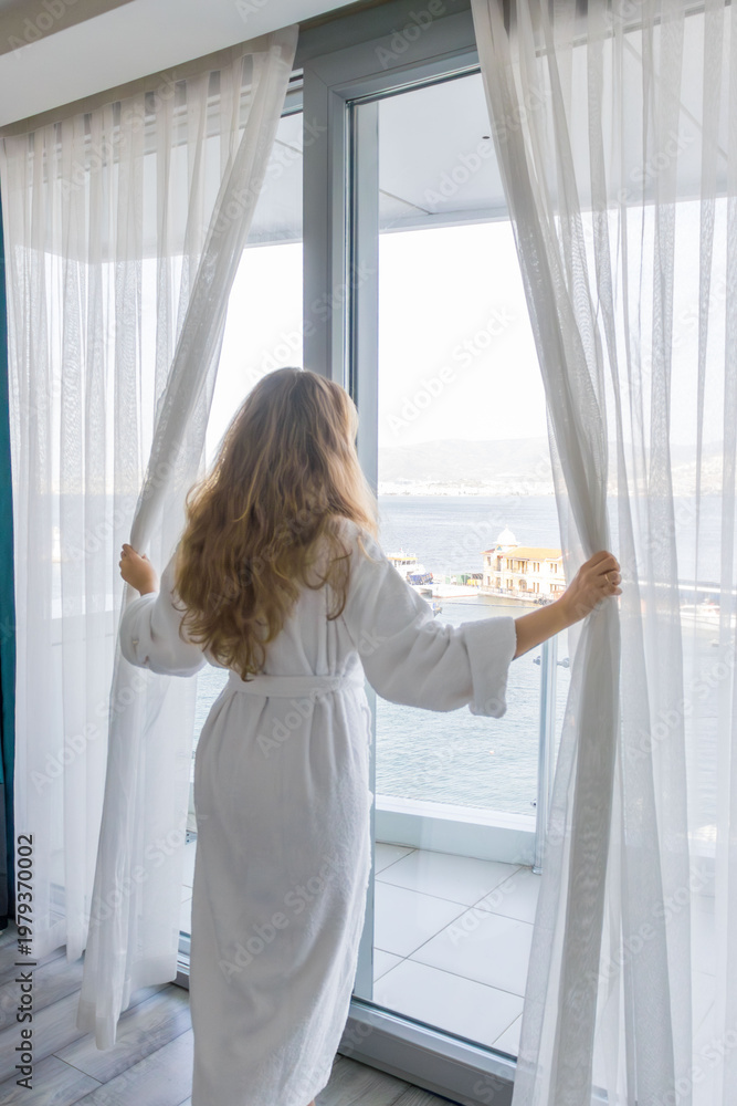 Fototapeta premium Woman opening curtains in apartment near large window with sea view. Morning routine, home lifestyle, natural light, daily life moment indoors