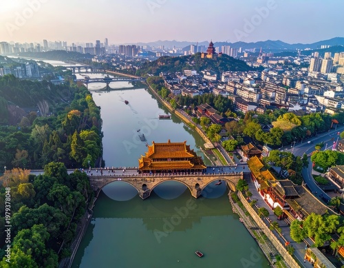 An aerial view showcases the historic Longmen Bridge gracefully spanning a tranquil river, framed by a sprawling cityscape and lush green hills in the background.