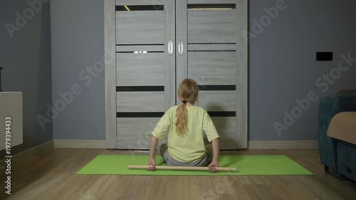 Back view of young girl with blonde hair in ponytail sitting on green yoga mat on wooden floor. She holds wooden stick, performing exercise for physical fitness indoors.