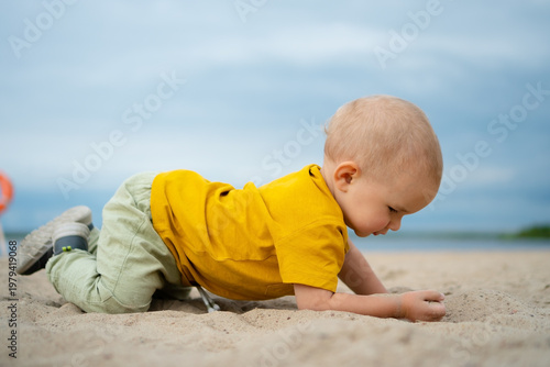 A happy toddler in a yellow shirt discovers the texture of sand on a bright beach day, embodying the wonder and excitement of exploring nature playground.
