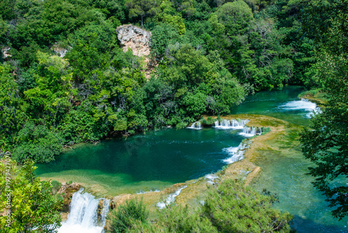 Krka National Park is of Croatia's natural treasures. Tranquil river surrounded by dense green trees, featuring small cascading waterfalls flowing gently, terraced rocks into clear green water