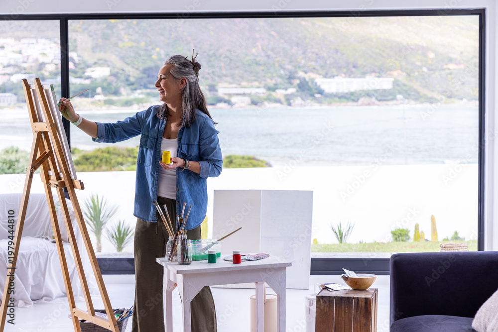 Naklejka premium Senior woman painting at easel in coastal home wearing denim with yellow paint jar, copy space