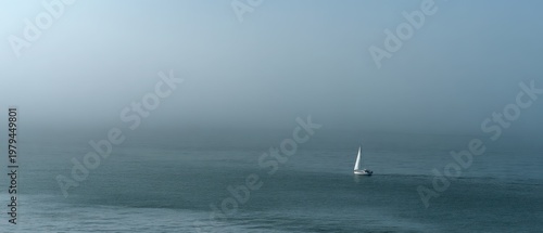 Aerial drone view of single white sailboat barely visible on vast misty teal ocean with overcast sky