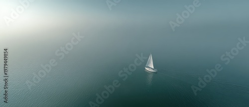 Aerial drone view of tiny white sailboat on glassy calm ocean with soft pastel mist and pale horizon