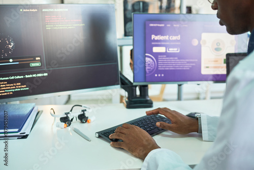 Photos African American male doctor typing at workstation, reviewing patient card interface and medical analysis data