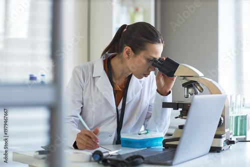 Canvastavla Young adult Caucasian woman scientist examining sample through microscope and taking notes in laboratory