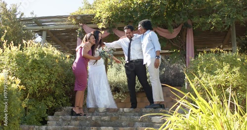 Wedding party huddling under pergola, linking and guiding bride in gown holding bouquet down steps