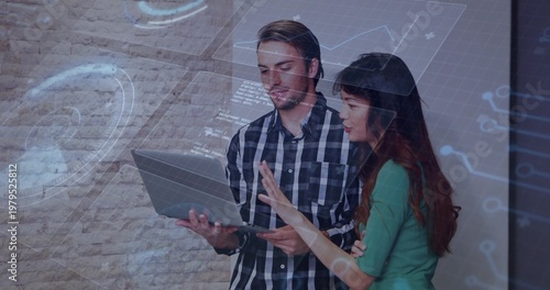 Colleagues examining laptop and pointing at screen in collaborative workspace with digital overlay
