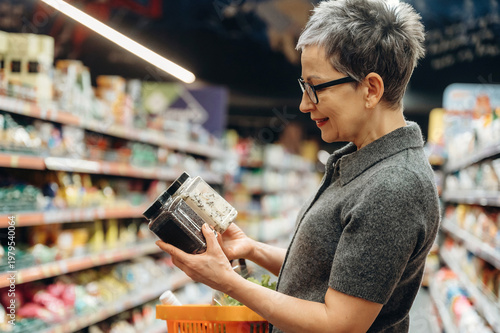 Mature woman with short gray hair examines spice jars while shopping in a grocery store aisle, colorful products displayed on shelves in the background