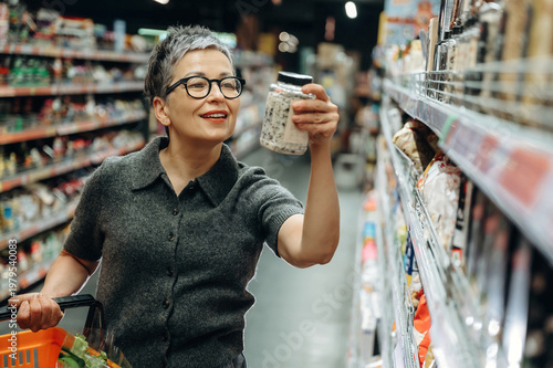 Middle-aged woman with short gray hair wearing glasses examines a jar of food while shopping in a grocery store aisle filled with various products and shelves