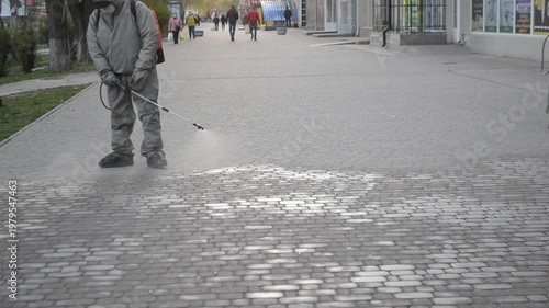 Man in airtight suit sprays disinfecting liquid chemicals antiseptic on street in city on day. Sanitation workers clean pavement, crosswalk. Sanitary measures in public place. Coronavirus pandemic
