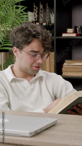 Young Caucasian male reading a book while seated at a desk with a laptop, colorful stationery, and bookshelves filled with books in a cozy indoor setting