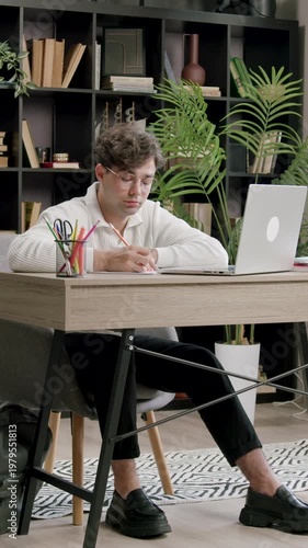 Young male student in casual attire sits at a desk with a laptop, focused on studying in a cozy home office surrounded by books and plants