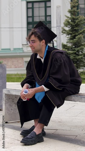 Male graduate in black cap and gown sits on bench, holding diploma and water bottle, with modern building and greenery in the background