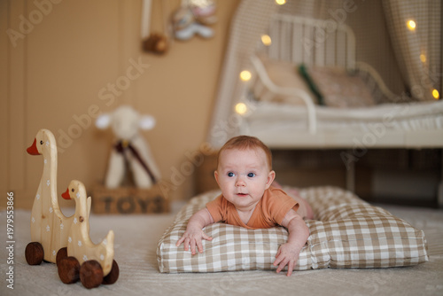 A baby lies on a checkered pillow on the floor near a wooden toy goose. Shallow depth of field