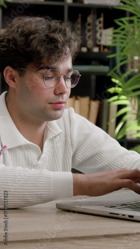 Young male student with curly hair wearing glasses types on laptop, showing frustration while seated at a desk with stationery and books in a cozy study environment
