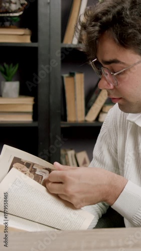 Young male reader in white sweater examines pages of an open book while seated at a wooden table in a cozy library filled with books and plants