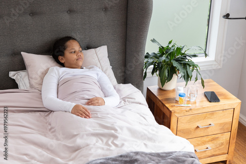 Child lying on pink duvet in bedroom, resting, looking at nightstand with pill-bottle and phone