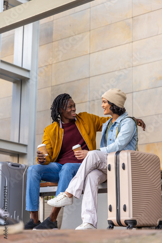 Diverse couple sitting on bench holding coffee cups at station concourse with beige suitcase