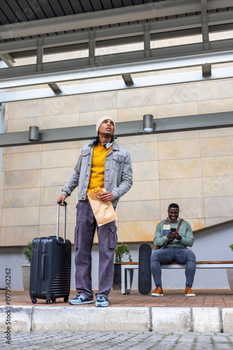 Standing traveler holding paper bag, pulling roller suitcase on station, denim jacket and headset