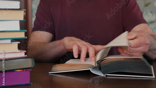 Close-up of a man's hands flipping through pages of a thick book at a wooden desk with a stack of books on the side.