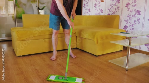 A man cleaning a laminate wooden floor with a green microfiber mop in a bright living room. Concept of domestic chores, home maintenance, hygiene, and daily housekeeping routi