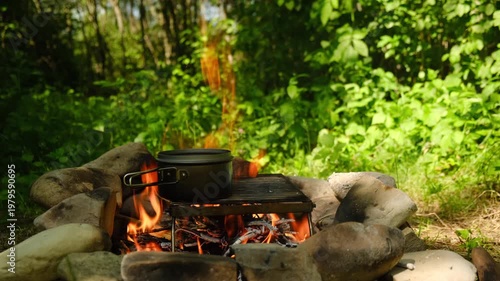 Male hand puts boiler with water over bonfire near green forest at summertime. Campfire cooking in spring season. Concept of adventure, travel and hiking. Fast dinner camping
