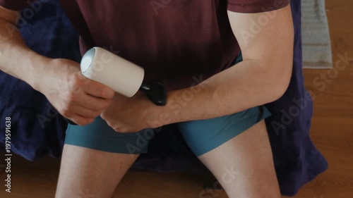 High-angle close-up of a man using a handheld electric massage gun on his forearm muscle