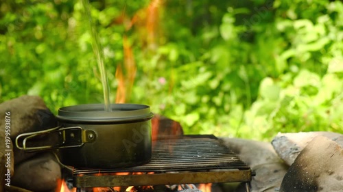 Close-up of clear fresh water being poured in metal pot on campfire at summer time. Peaceful and serene outdoor scene. Camping concept