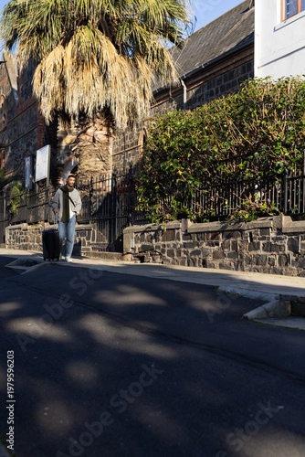 Man 20s walking on sidewalk in gray jacket pulling rolling suitcase by palm tree, copy space