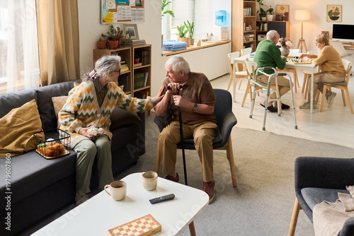 Bild auf Leinwand Wide shot of senior residents socializing in care home, woman comforting man with cane while couple playing board game in background