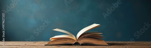 Open book rests on rustic wooden table against dark blue backdrop. Pages flutter slightly, inviting readers to explore knowledge, stories, or academic pursuits. Copy space available.