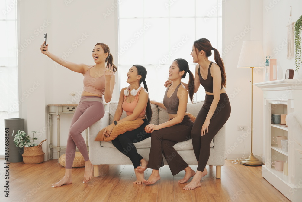 Obraz premium A group of four beautiful Asian women in sports bras meet and greet each other, then sit and chat on a sofa and take selfies together at the studio. The concept of exercising for health is reflected i