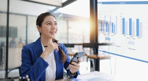 A professional Asian female executive sitting at a desk, looking at a data visualizations, graphs, and business analytics. She is holding a smartphone and has printed reports on the table.
