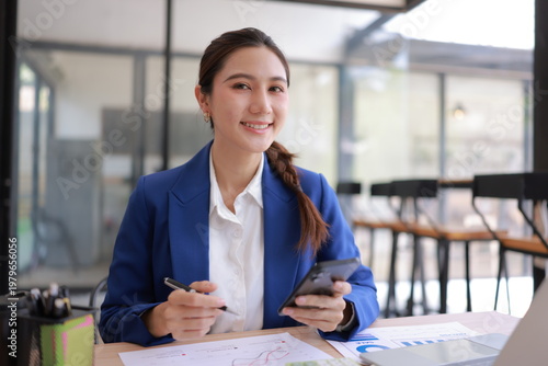 A smiling young Asian female entrepreneur sitting at a desk, analyzing real estate business data and financial graphs on a laptop and paper documents. She is holding a pen, appearing professional and 