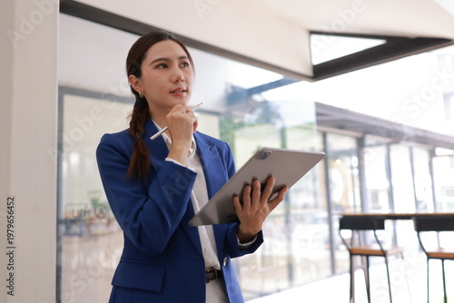 Confident young Asian businesswoman in blue suit using digital tablet with stylus pen in modern office background.