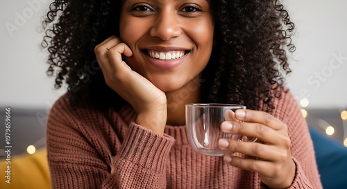 A young woman with curly hair smiles warmly while holding a clear mug of a beverage.