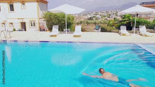 Man swimming in a clear outdoor pool viewed from drone. Sun sparkles, ripples and soft umbrella motion create a peaceful, warm and refreshing summer scene.