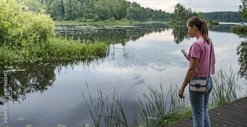 Wallpaper Mural Young woman stands by calm lake, holding phone Torontodigital.ca