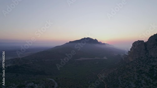 Drone shot of a mountain range at sunset with the sun disappearing behind the horizon. Golden haze and soft light create a peaceful, minimal and atmospheric scene.
