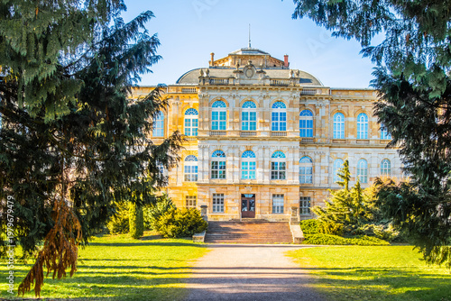 Herzogliches Museum Gotha centered in landscaped park, historic palace facade framed by evergreen trees, green lawn and straight path under clear blue sky, cultural museum landmark in Thuringia