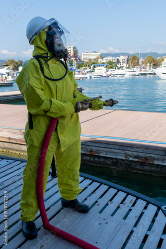 Fire drill on yacht stern: crew member in protective suit uses fire hose and breathing apparatus during training, simulating emergency firefighting on board.